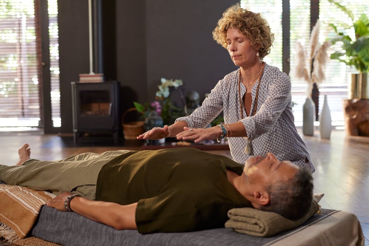 Woman performing reiki energy healing on man at wellness center. Holistic therapist holding hands above body client to restore balance and calm.