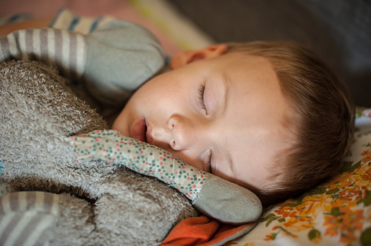 A young child lies asleep on a colorful blanket, holding a soft stuffed toy